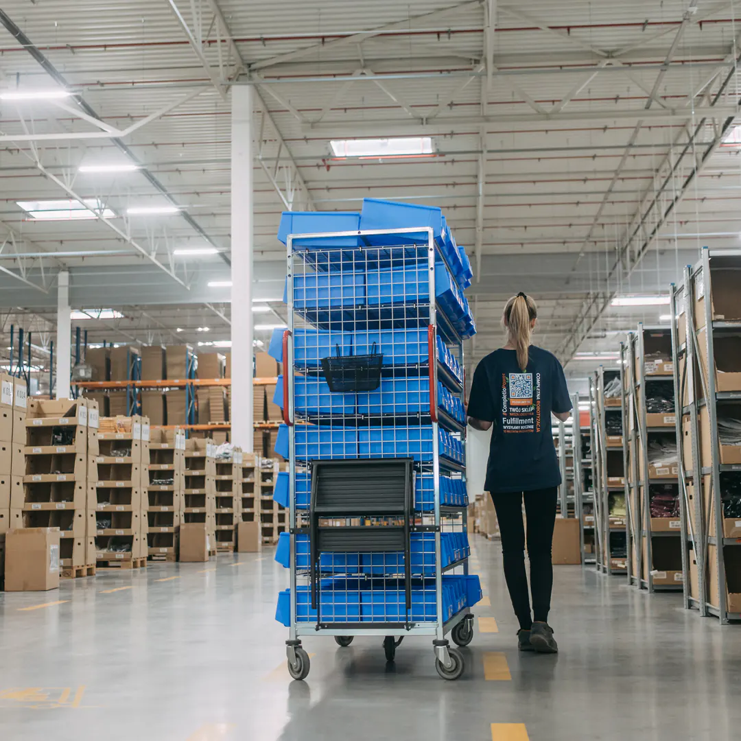 A warehouse worker wheels a trolley of blue totes down an aisle between racks of received goods